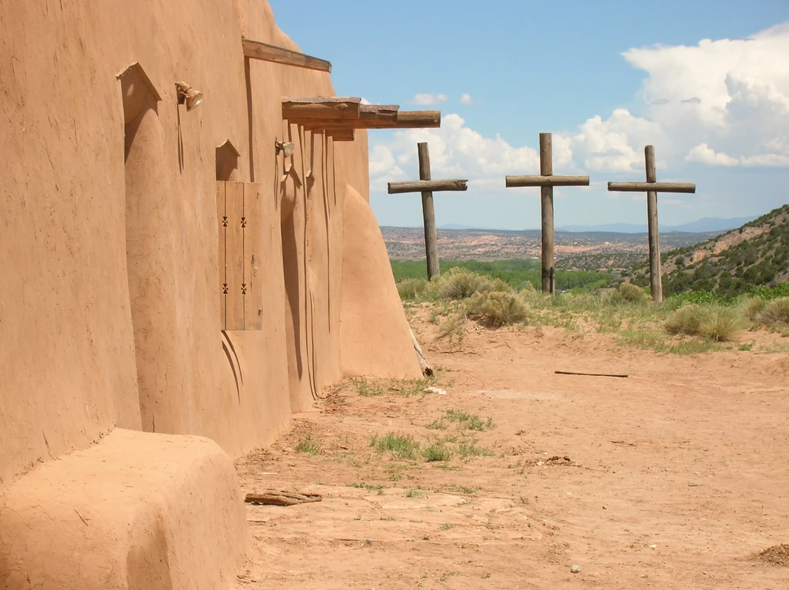 A view of a light brown mud-plastered wall of an adobe chapel with three tall wooden crosses just beyond it and hills and sky in the distance.