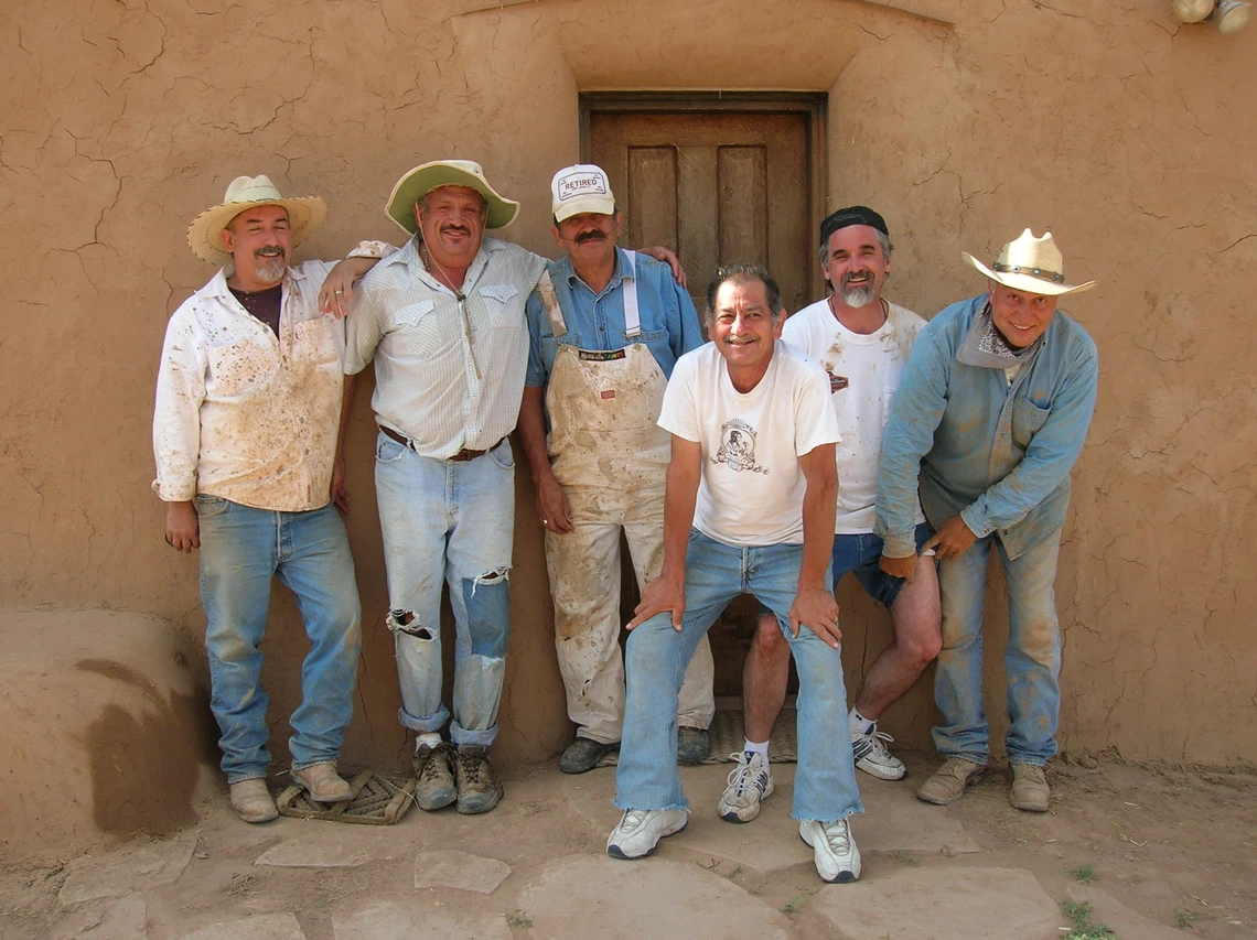A group of men with smiles on their faces pose in front of the doorway of an adobe chapel freshly re-plastered with mud.