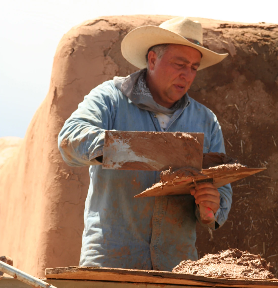 A man wearing a blue shirt and cowboy hat uses a trowel to spread a wet mud mixture on the wall of an adobe chapel.