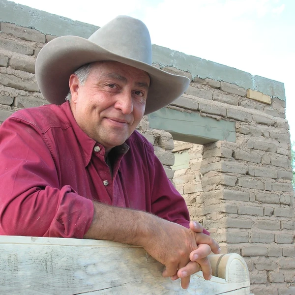 A man wearing a cowboy hat and red shirt stands in front of an adobe brick structure.