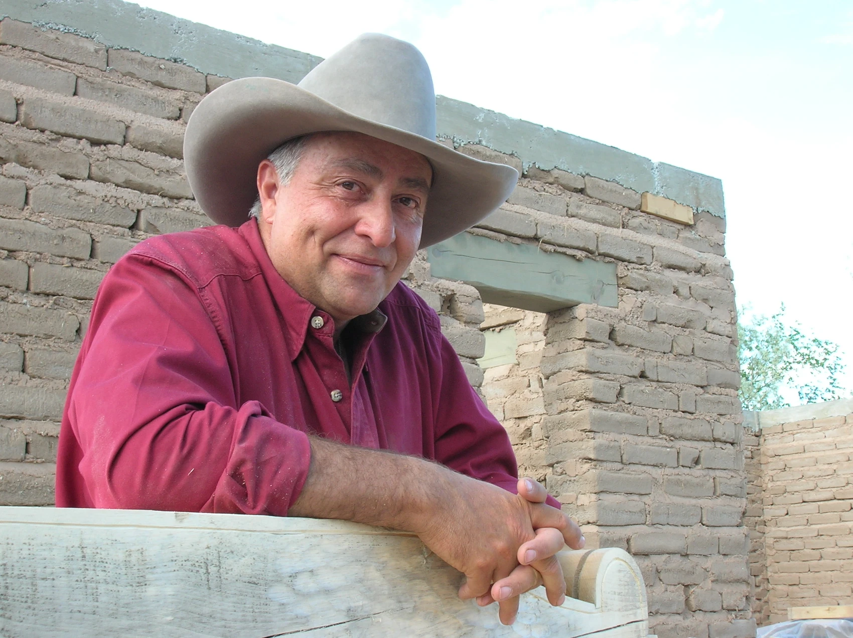 A man wearing a cowboy hat and red shirt stands in front of an adobe brick structure.