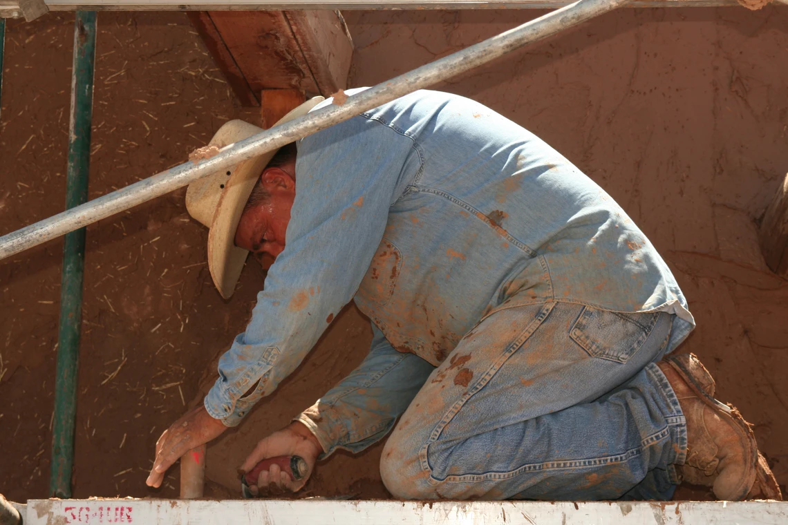 A man wearing a blue shirt and cowboy hat bends down on his knees on scaffolding to use a trowel to spread a wet mud mixture on the wall of an adobe chapel.