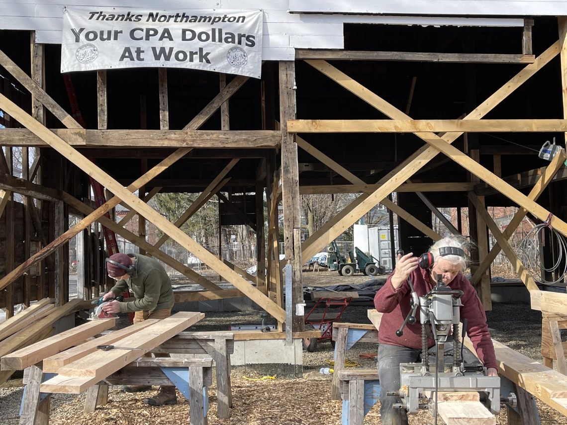 A woman at an outdoor construction site straddles a long plank of wood mounted on sawhorses while drilling holes with a hand-powered metal drilling tool; large wooden vertical and horizontal beams form the skeleton of a timber-frame barn behind her.