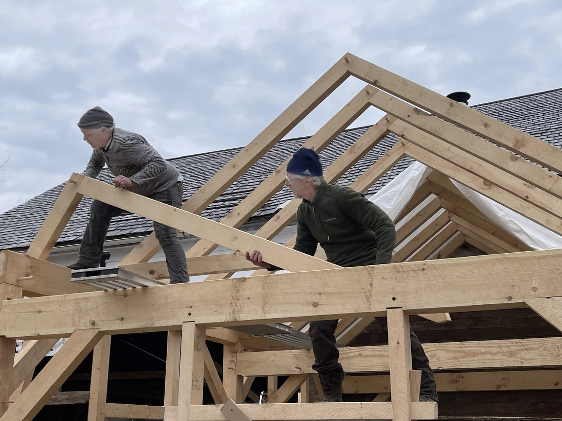 A woman and man work on the top of a large wooden beam on a roof to position a triangular timber-frame truss in place.