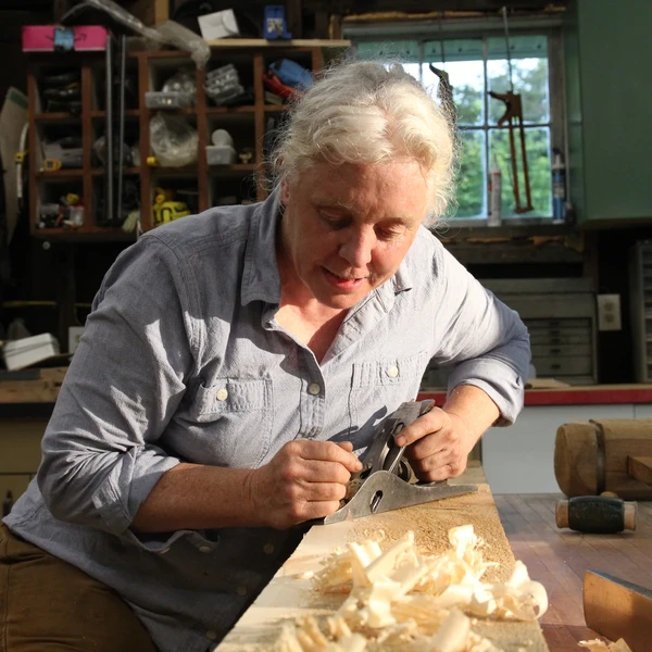 A woman in a light blue shirt uses a plane to smooth out a piece of wood on a worktable in her workshop.