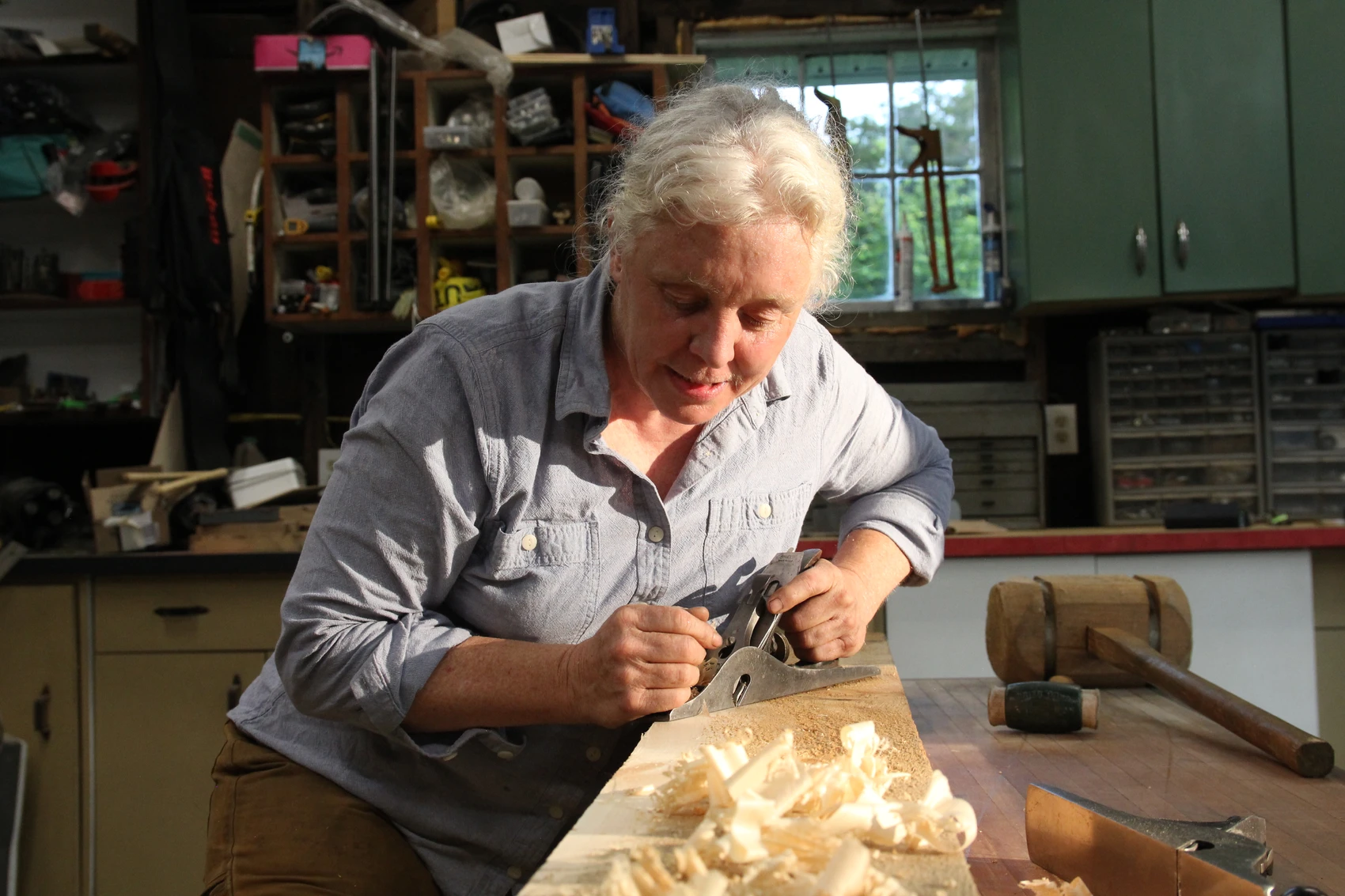 A woman in a light blue shirt uses a plane to smooth out a piece of wood on a worktable in her workshop.