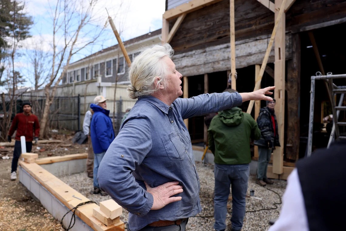 A woman in a blue shirt points her arm to direct a group of volunteers where to place a wooden beam at an outdoor construction site for the restoration of a timber-frame barn.