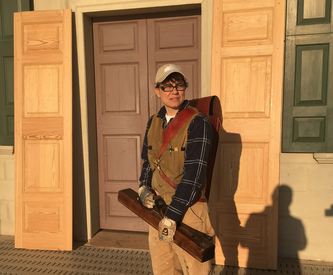 A woman in work clothes and holding a narrow brown leather tool case poses in front of an old wooden door flanked on either side by a pair of new wooden replacement doors.