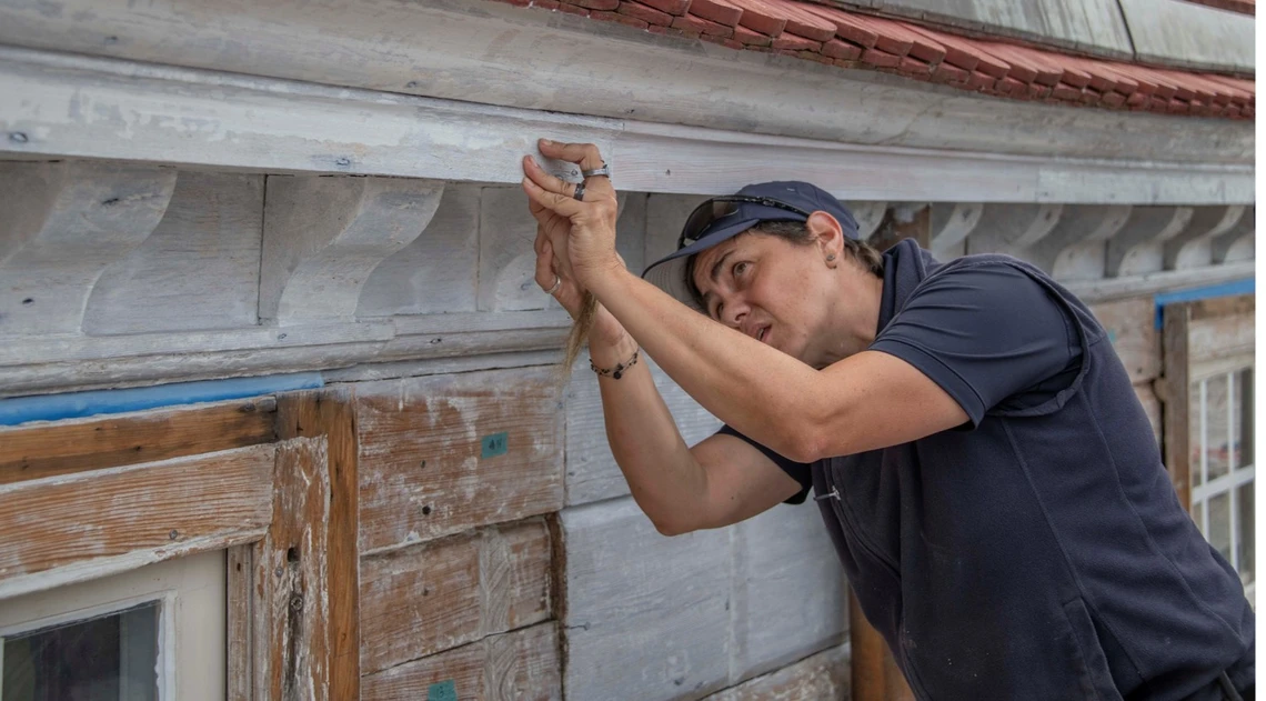 A woman wearing a navy blue shirt and cap carefully examines an old, white-painted strip of wood that is running along the length of the eaves of an historic house.