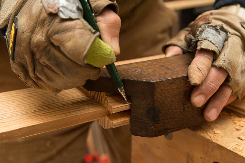 A close-up image of a woman’s hands wearing brown open-fingered gloves as she marks a small piece of wood with a pencil.