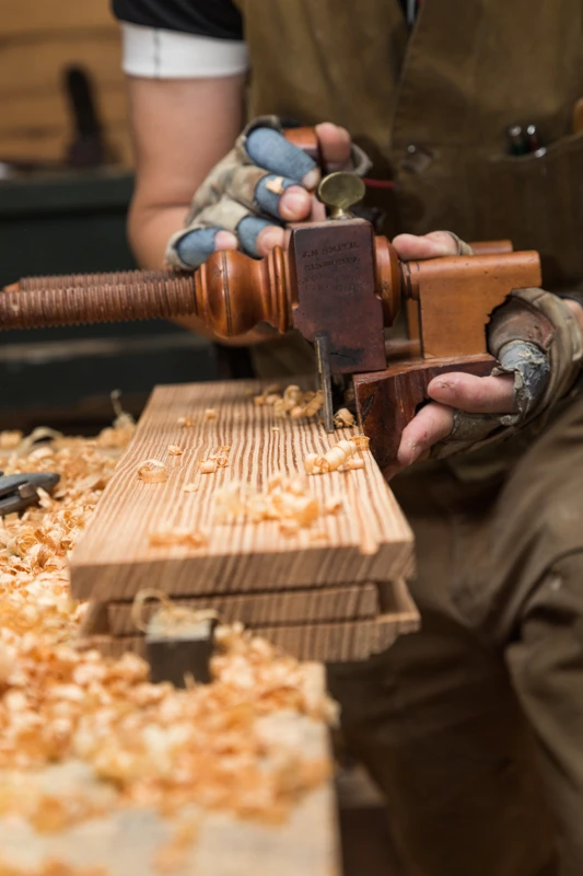 A close-up image of a woman in brown work clothes cutting a groove in a plank of wood with an old, historic wooden tool at a worktable covered with golden-colored wood shavings.