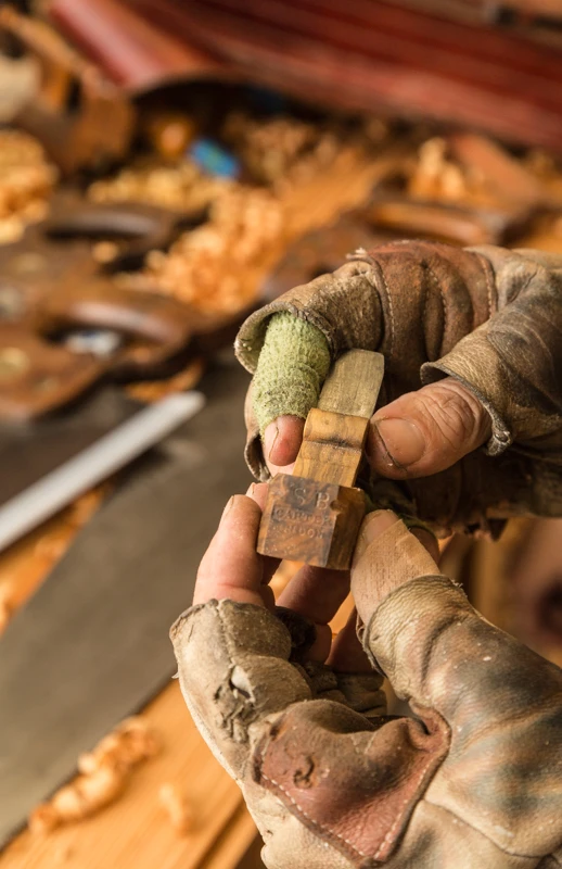 A close-up image of a woman’s hands wearing brown open-fingered gloves and holding a small piece of wood above her worktable; the table is topped with golden wood shavings and planks of wood.