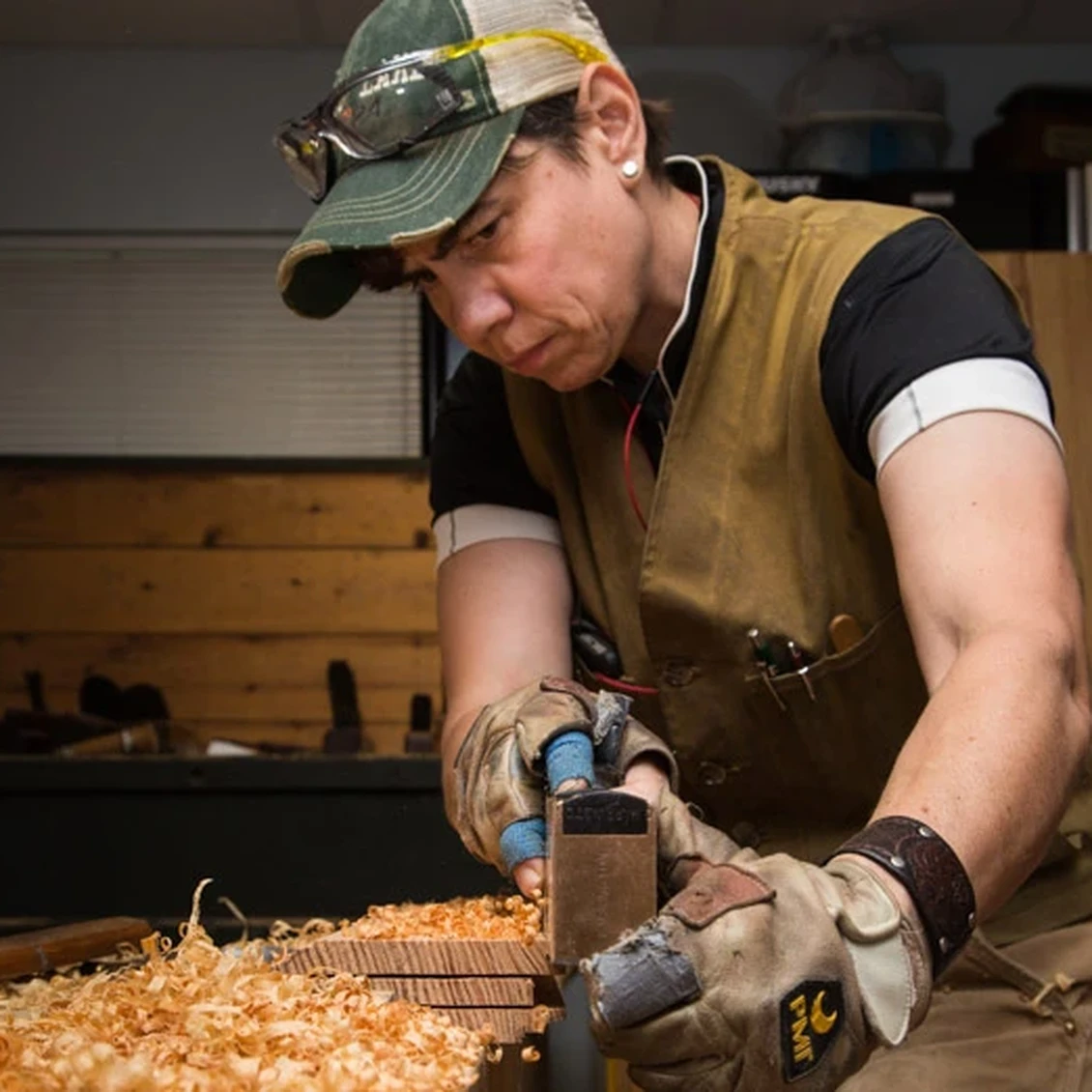 A woman wearing a cap, brown vest, and work gloves runs a plane on a piece of wood on a worktable in her workshop, which is topped with a small pile of light brown wood shavings.