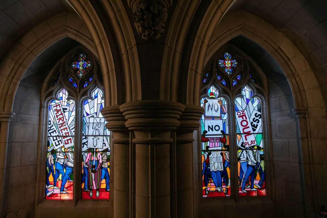 Two brightly colored vertical stained-glass windows set in the limestone moldings of a cathedral wall feature groups of people marching and carrying signs that say “Fairness” and “No Foul Play.”