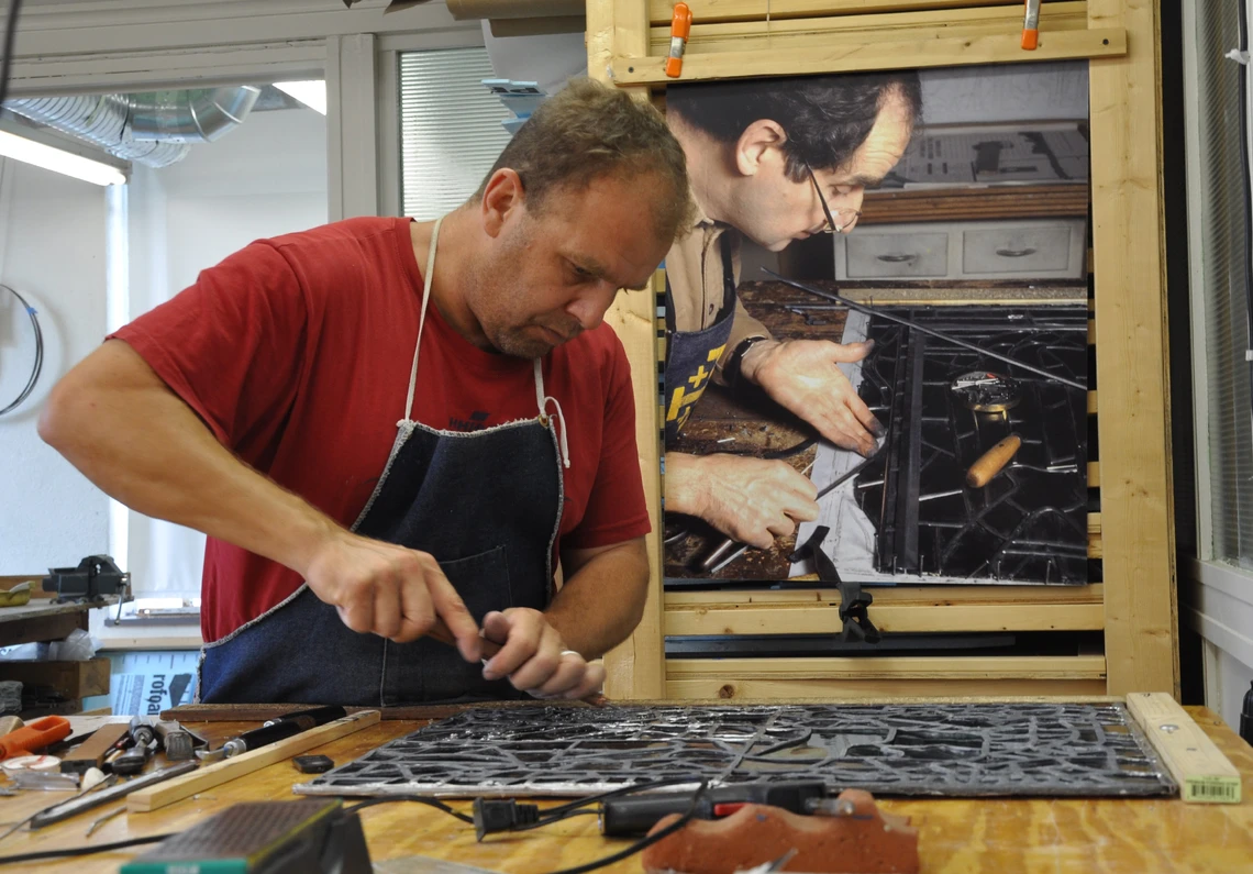 A man in a red t-shirt and dark blue apron works with a sharp metal tool to cut the thin lead cames of a stained-glass window that he is repairing on a wooden worktable in his studio.