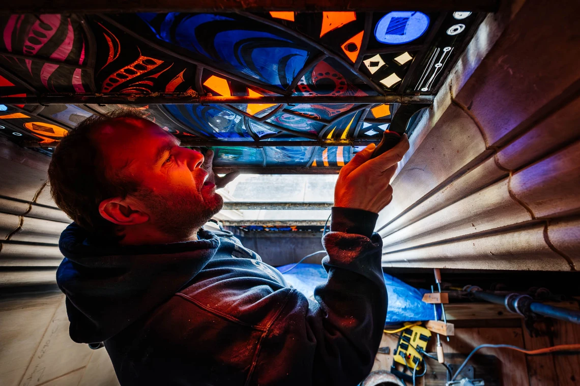 A man in a dark blue sweatshirt uses a metal tool to install a multi-colored stained-glass window in an opening in the limestone wall of a cathedral; red-colored light shines on his face from the reflection of sun through the window.