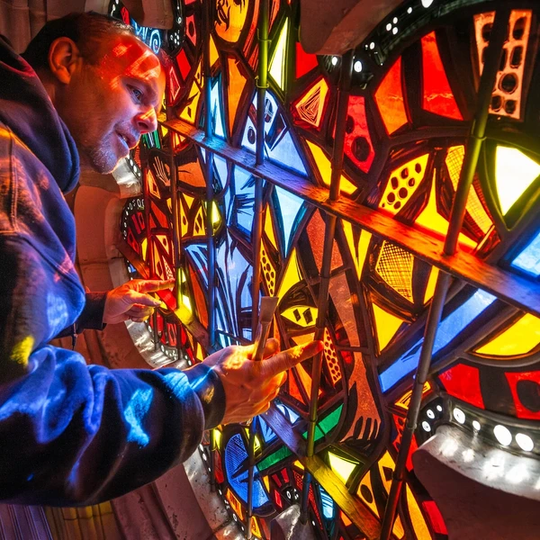 A man works on a stained-glass window that features a mosaic of bright yellow, orange, red, and blue pieces of glass located in the architectural opening of a limestone wall in a cathedral; red-colored light shines on his face and sweatshirt from the reflection of the sun streaming through the window.