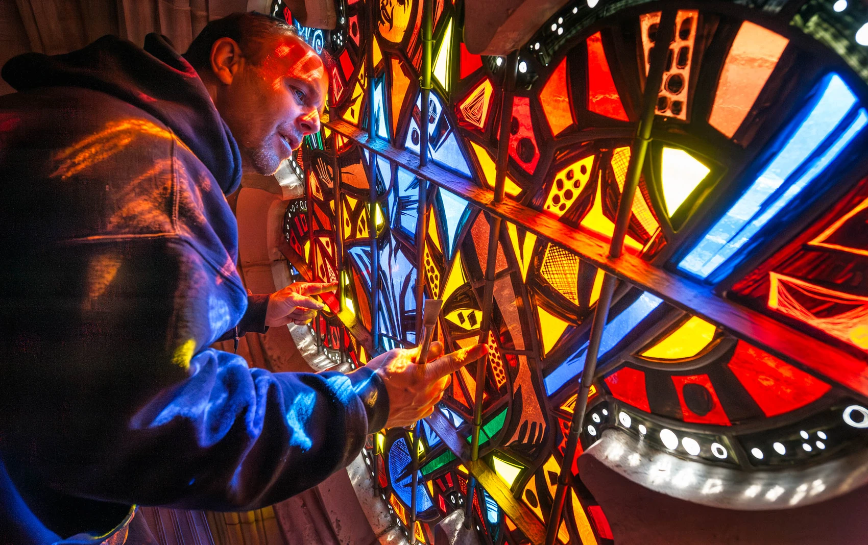 A man works on a stained-glass window that features a mosaic of bright yellow, orange, red, and blue pieces of glass located in the architectural opening of a limestone wall in a cathedral; red-colored light shines on his face and sweatshirt from the reflection of the sun streaming through the window.