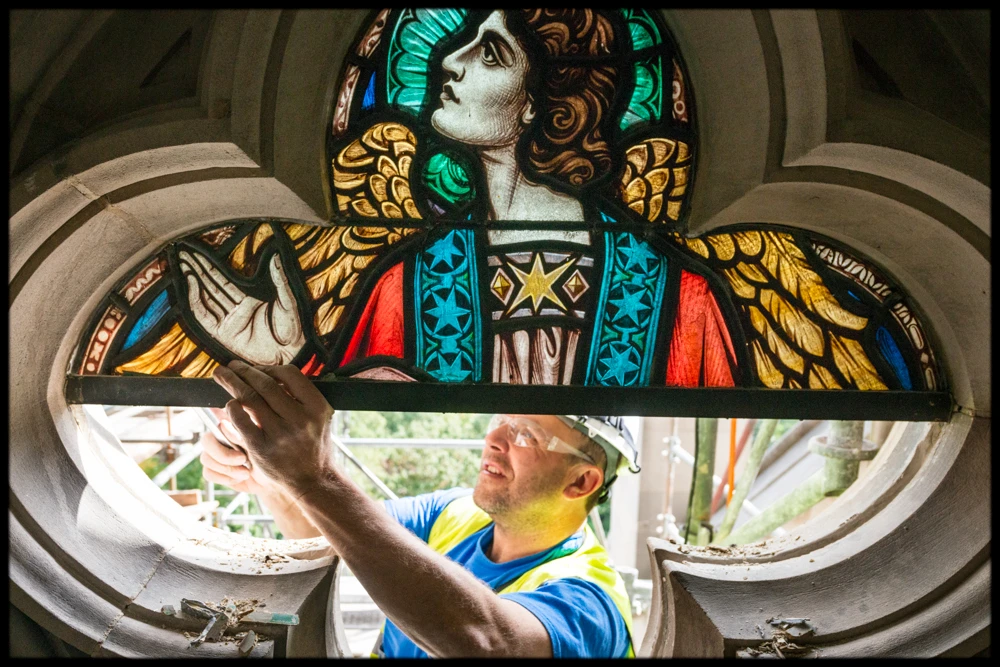 A man in a hard hat reaches up to install a brightly colored stained-glass window featuring the image of an angel into a quatrefoil-shaped architectural opening in the limestone wall of a cathedral.