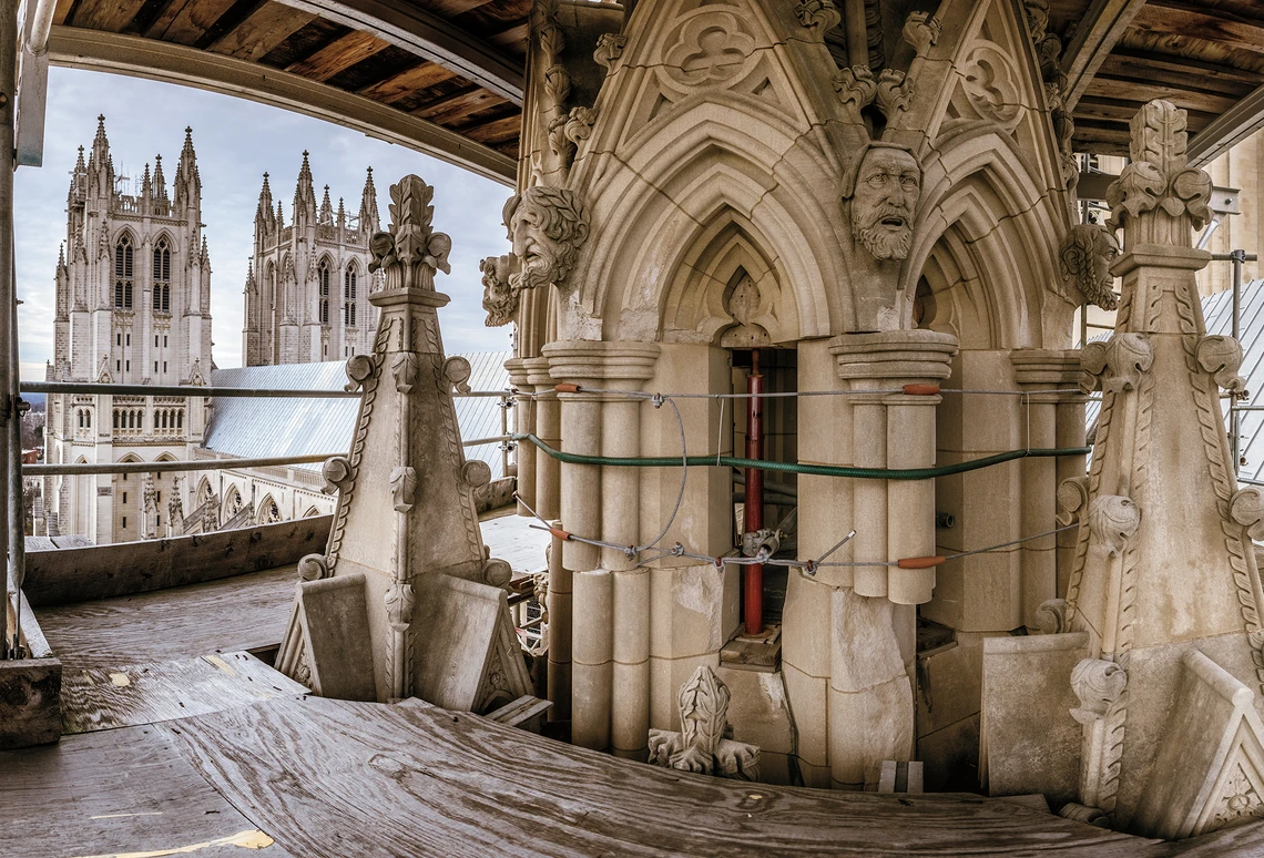 A panoramic view of damaged limestone columns and carvings of a giant Gothic-style pinnacle, which is held together by three stabilization cables in scaffolding near the top of an exterior tower of a cathedral.