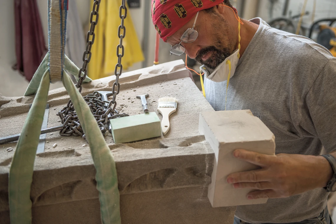 A stone carver wearing a red bandana on his head fits a small square limestone block into the bottom corner of a damaged Gothic-style ornamental limestone pinnacle stone that is lying sideways on a workbench in his workshop.