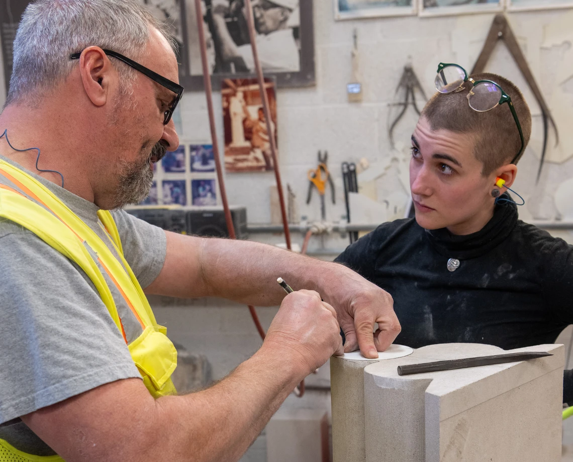 Inside a stone masons’ workshop, a man wearing a yellow safety vest shows a young woman how to trace the outlines of a cardboard template onto the top of a small limestone molding in preparation for carving.