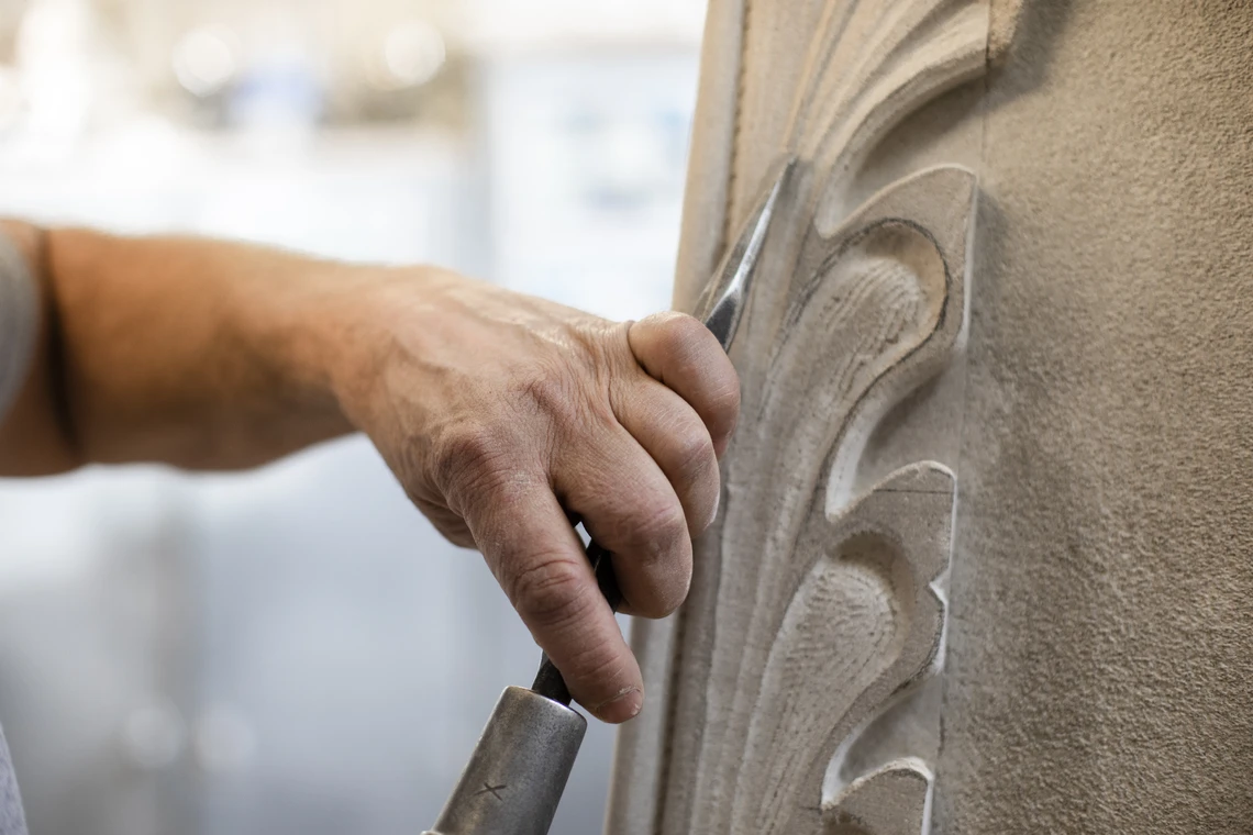 A close-up image of a man’s hand holding a metal chisel as he carves the veins of decorative foliage into a piece of limestone.