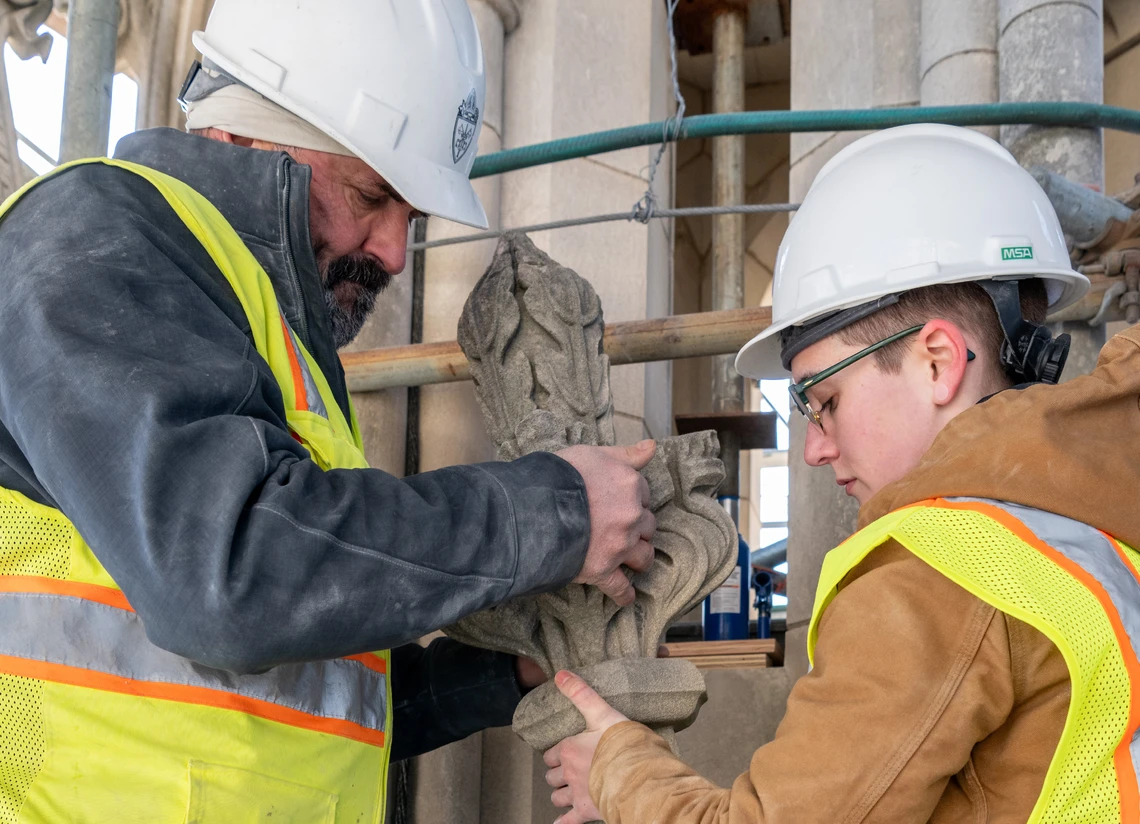 A man and a woman each wearing a white hard hat and yellow safety vest lift a small ornamental finial carving into place on the exterior of a Gothic-style cathedral.