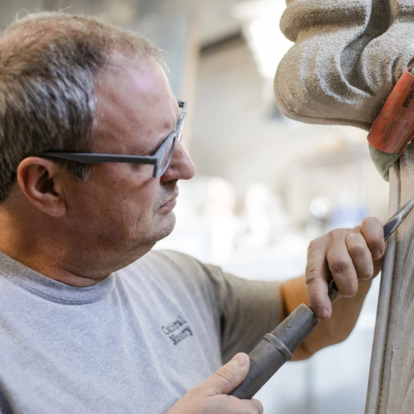 A man wearing glasses uses a metal pneumatic hammer and chisel to carve decorative foliage on a Gothic-style limestone pinnacle.