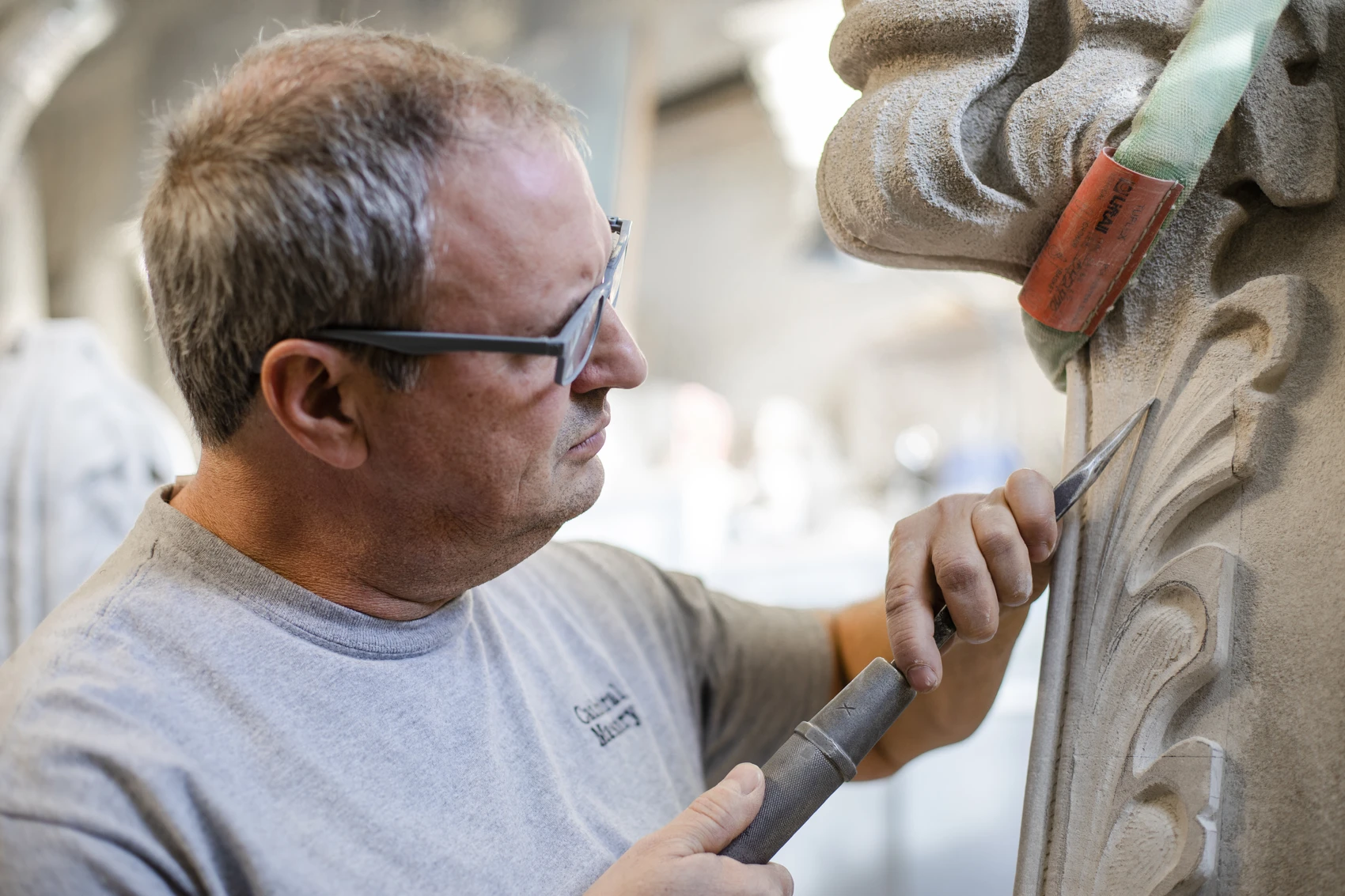 A man wearing glasses uses a metal pneumatic hammer and chisel to carve decorative foliage on a Gothic-style limestone pinnacle.