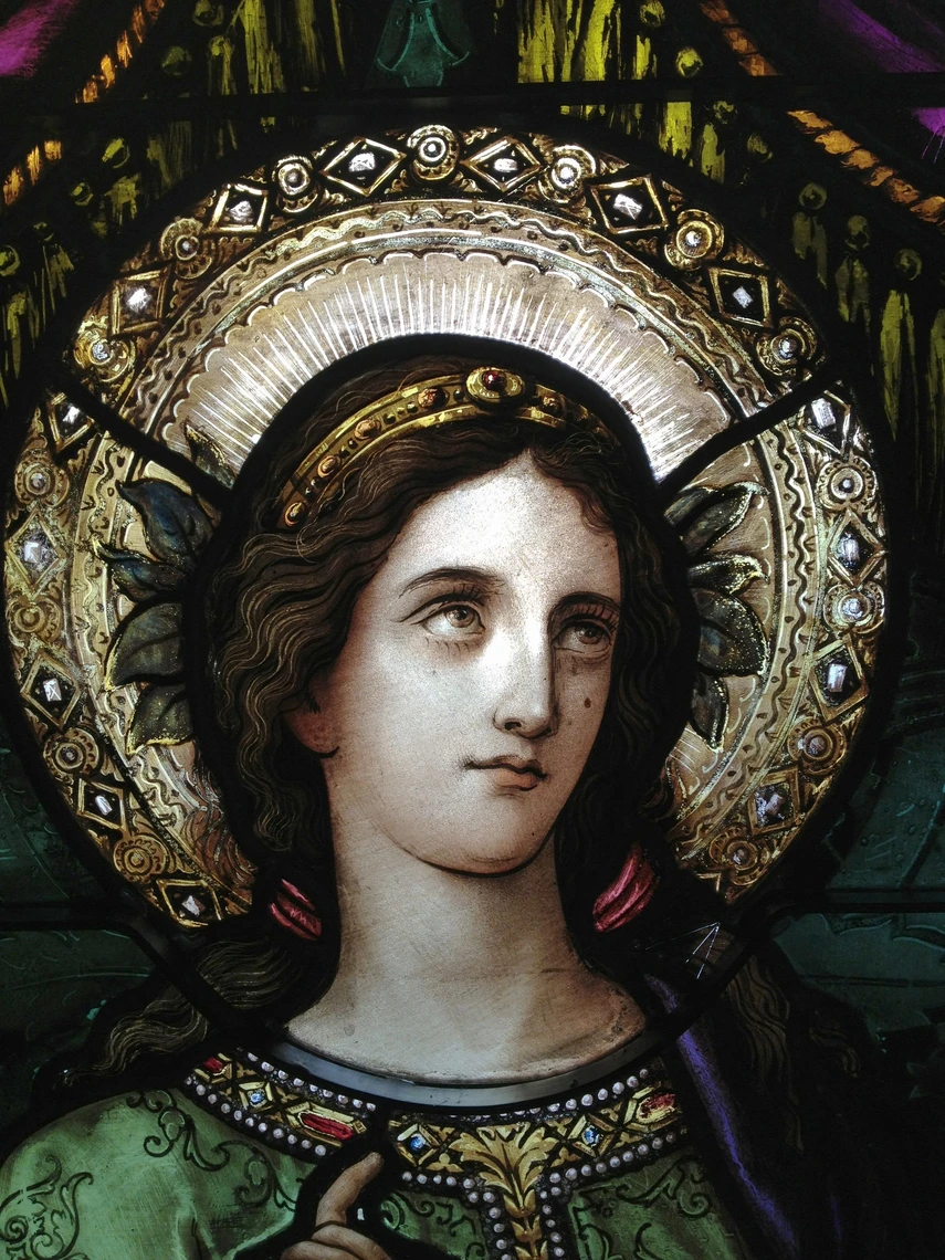 A detail of a stained-glass window features an image of the face of Saint Cecilia with long dark brown hair, a gold headband, and a silver-gold halo surrounding her head.