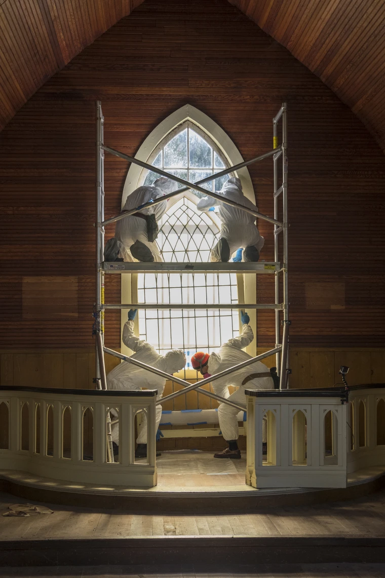 Four people dressed head to foot in white protective equipment work in scaffolding to remove a vertical arched window from an opening in the wall of an old church.