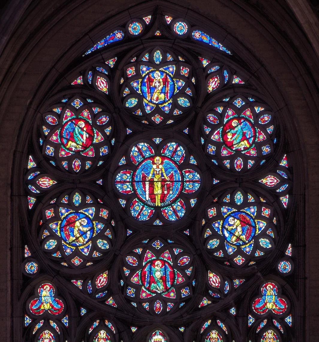 A large stained-glass window situated in the limestone tracery of a cathedral wall features images of saints and a kaleidoscope of bright colors, including red, yellow, and blue. 