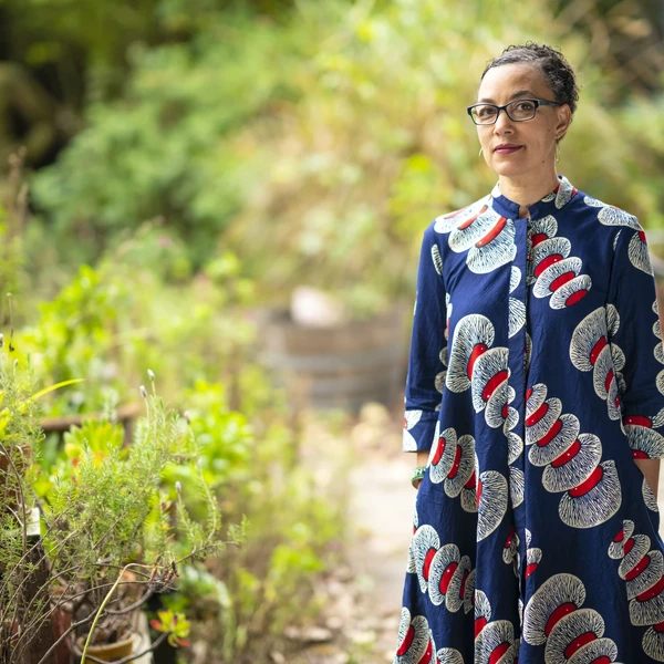 A woman with short dark hair and glasses wearing a dark blue dress patterned with red and white circular motifs stands in a lush garden surrounded by green bushes and trees.