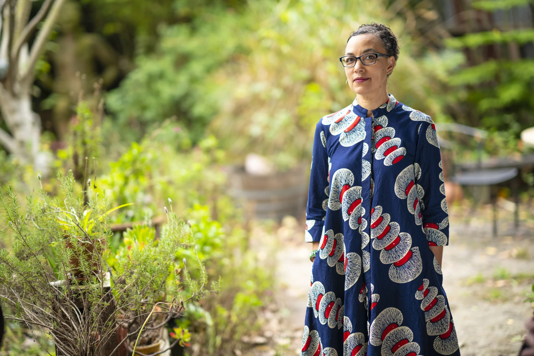 A woman with short dark hair and glasses wearing a dark blue dress patterned with red and white circular motifs stands in a lush garden surrounded by green bushes and trees.