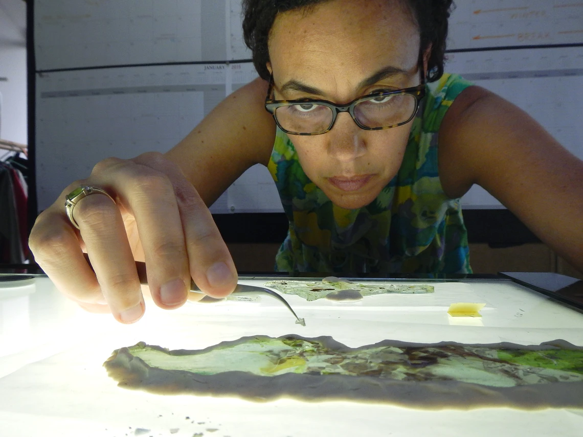 A close-up view of a woman with glasses holding a small metal tool in her right hand and working intently to repair pieces of glass on a light table.
