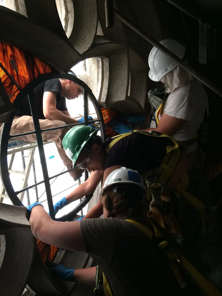Four people in hard hats and blue plastic gloves work to remove a window from a limestone opening in the stone wall of a church.