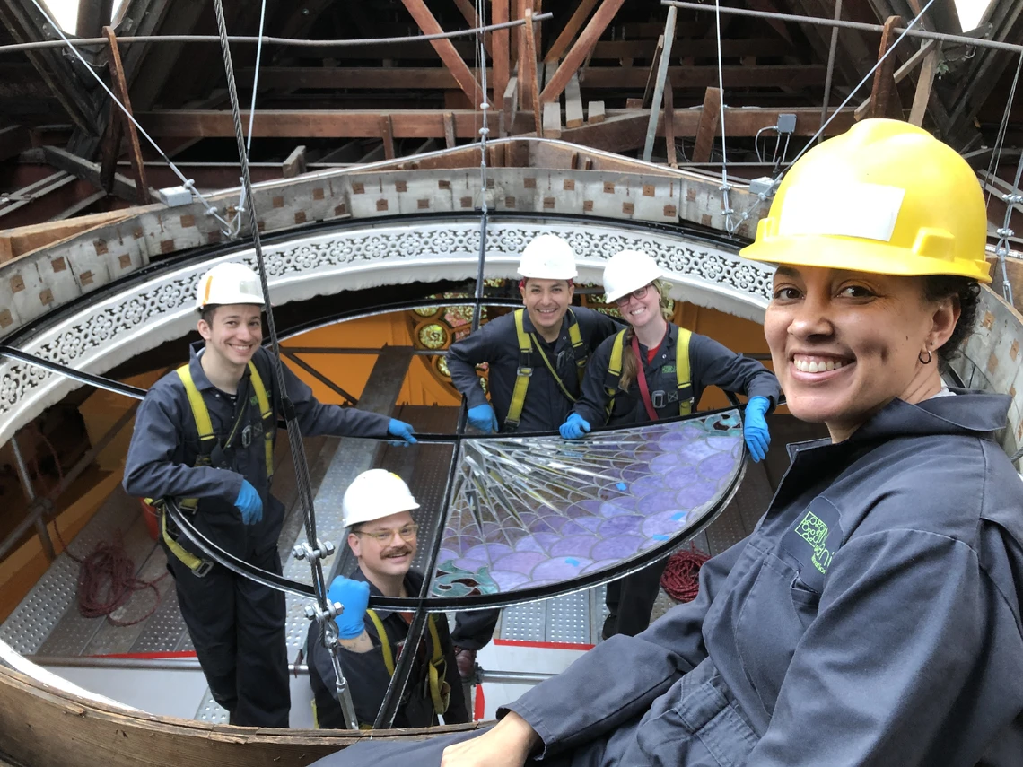 A group of five smiling people in hard hats pose in scaffolding in front of a large piece of stained glass that they are installing inside the circular dome opening of a building.
