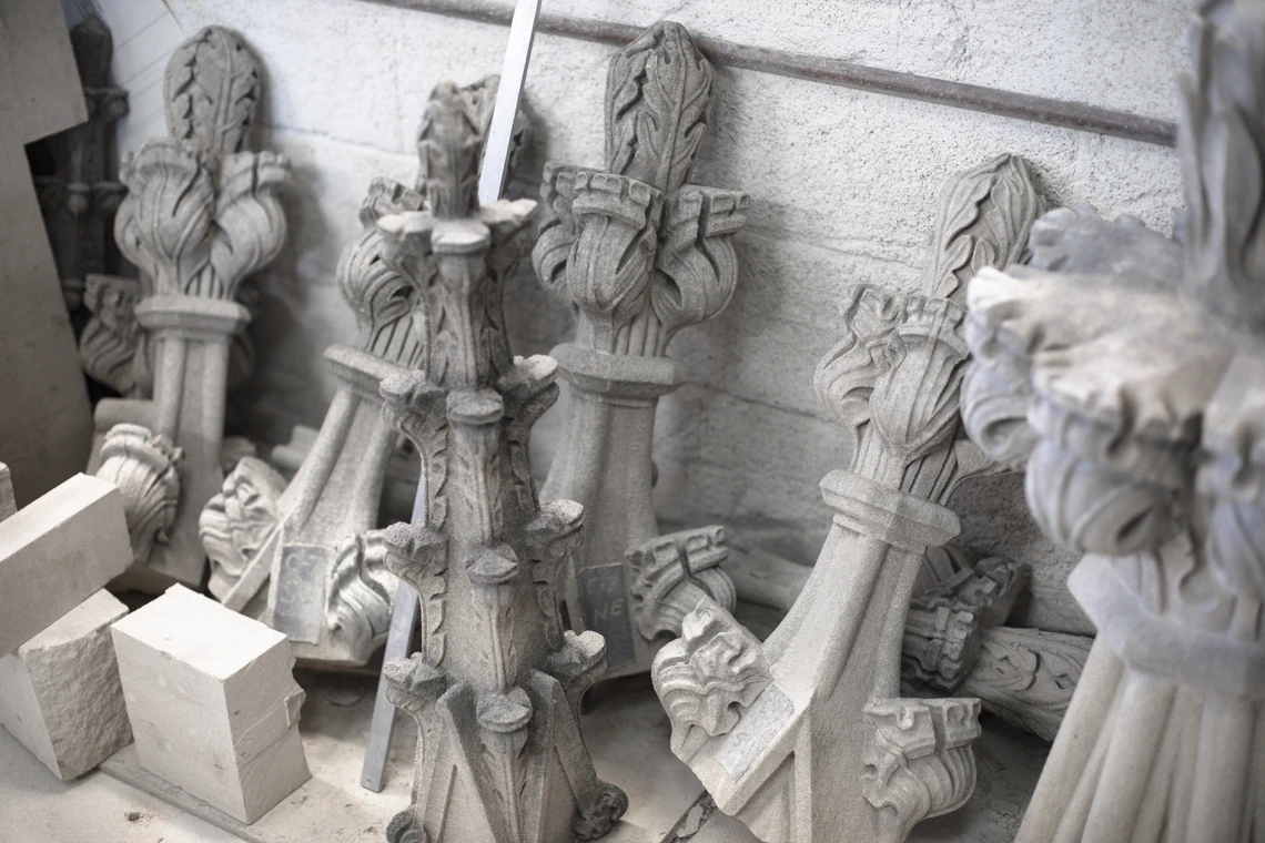 A row of six small Gothic-style limestone finials and three small square blocks of limestone lean against the wall of a cathedral workshop.