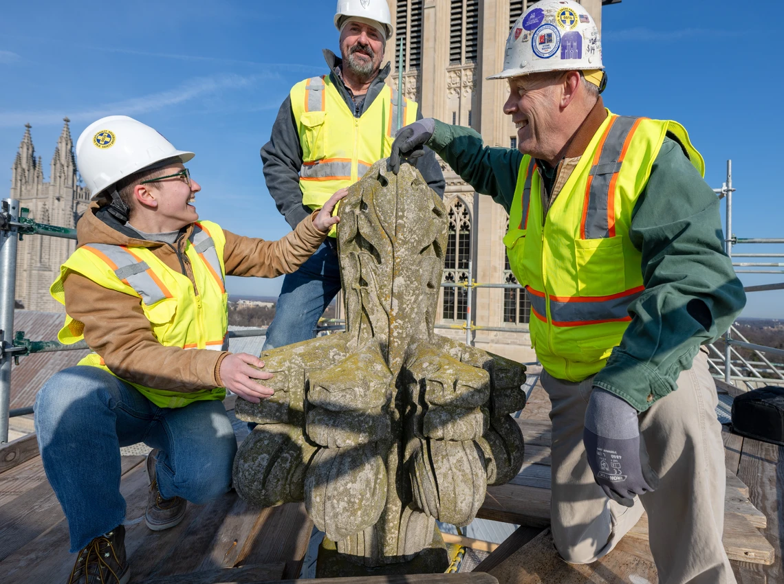 A young woman and two men in white hard hats and yellow safety vests pose around an old Gothic-style finial stone up high on an outdoor scaffolding platform. Two tall cathedral towers are behind them.