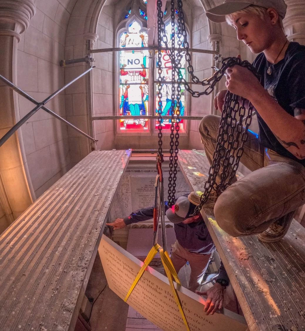 A young woman kneels on a scaffolding platform and pulls on a chain hoist to lift a rectangular limestone tablet into place inside a cathedral; a colorful stained-glass window is visible behind her.
