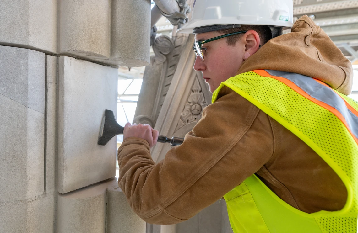 A young woman in a white hard hat and yellow safety vest uses a wide metal chisel to carve the flat surface of a limestone column on the exterior of a Gothic-style cathedral.