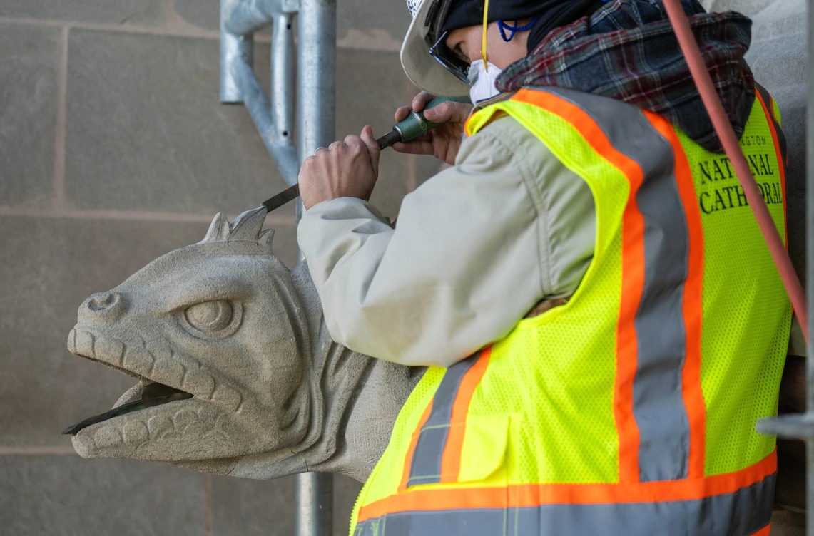 A young woman in a hard hat and yellow safety vest works up in scaffolding to carve an iguana-shaped gargoyle on the exterior of a cathedral.
