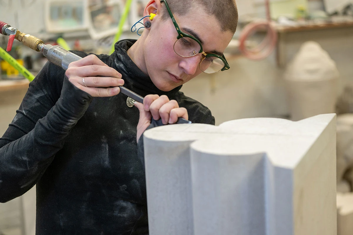 A young woman with short brown hair and glasses works inside a workshop to carve a curved piece of limestone molding by hand with a metal hammer and chisel.