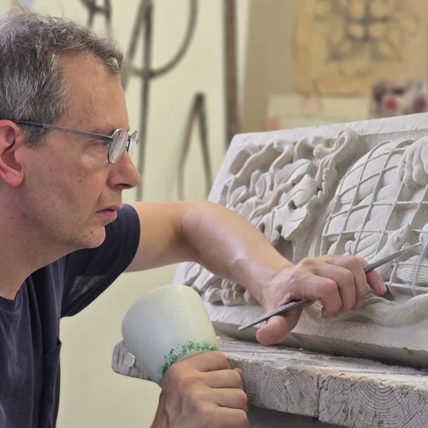 A man in a workshop uses a mallet and chisel to carve an intricate ornamental design into a rectangular piece of limestone on a wooden work bench.