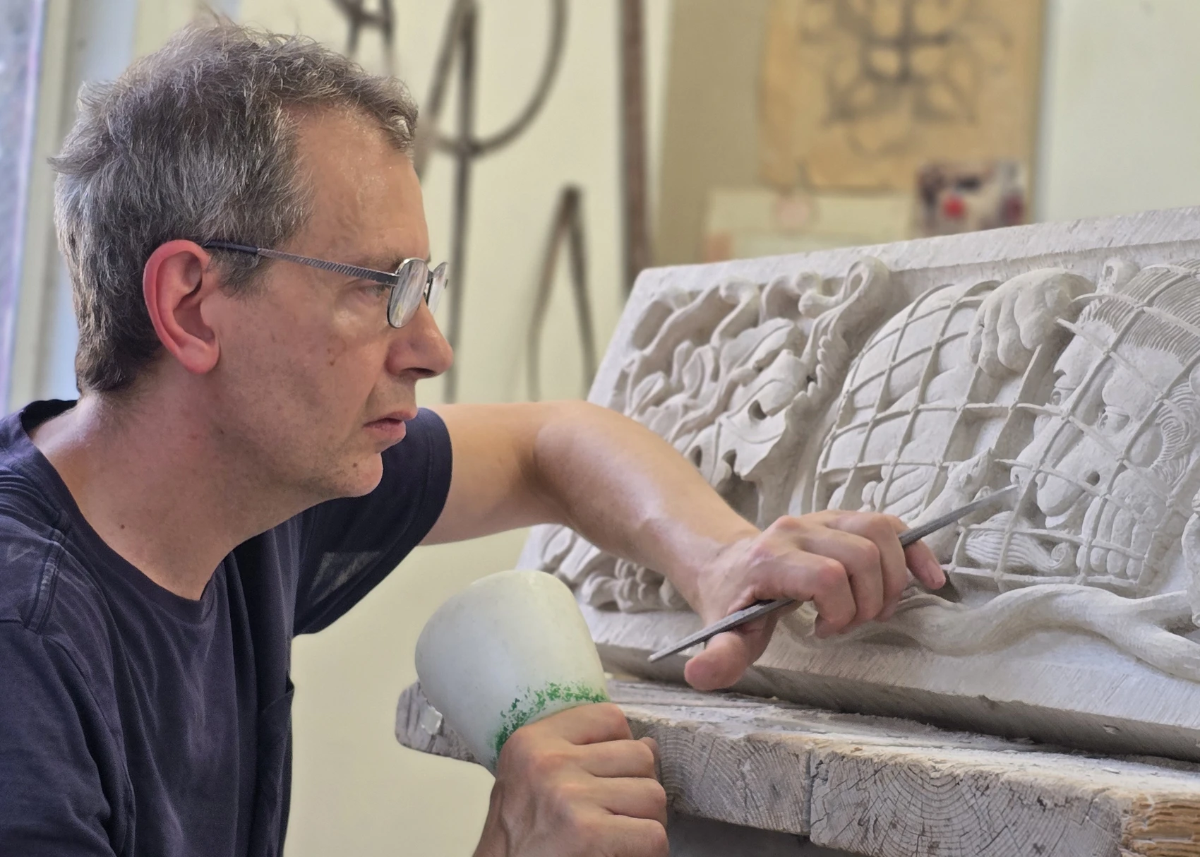 A man in a workshop uses a mallet and chisel to carve an intricate ornamental design into a rectangular piece of limestone on a wooden work bench.