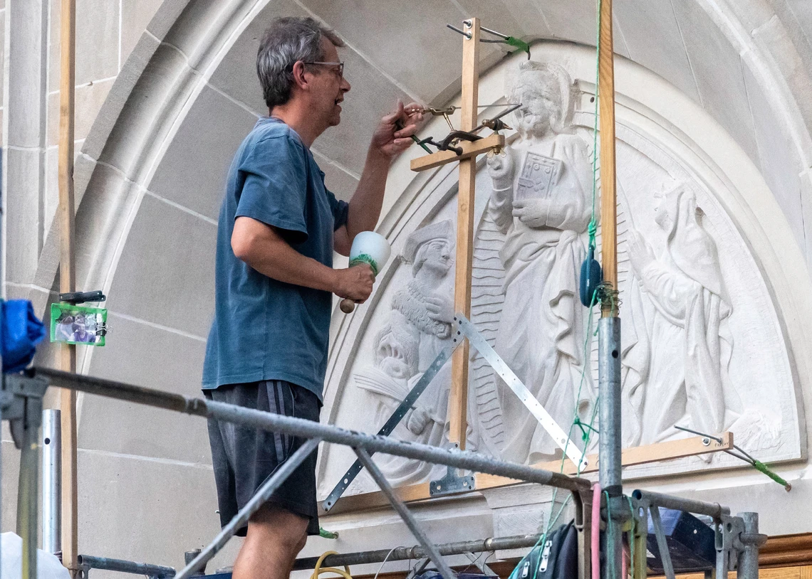 A man works in scaffolding on the exterior of a church to carve a large triangular sculpture depicting Jesus holding a bible flanked by two figures; the man is using a mallet and chisel and a metal measuring device to carve the figures in stone.