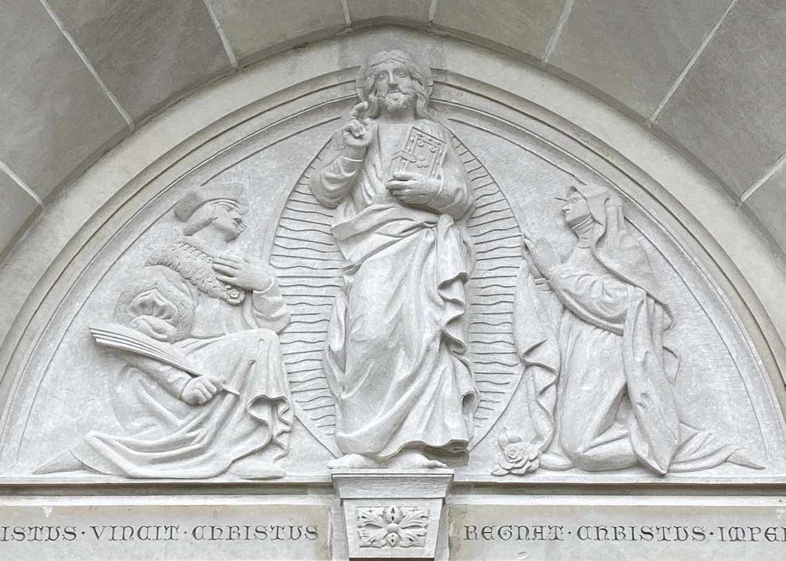 A triangular limestone tympanum sculpture located on the exterior wall of a church depicts a figure of Jesus holding a bible and two men kneeling on either side of him who are praying.