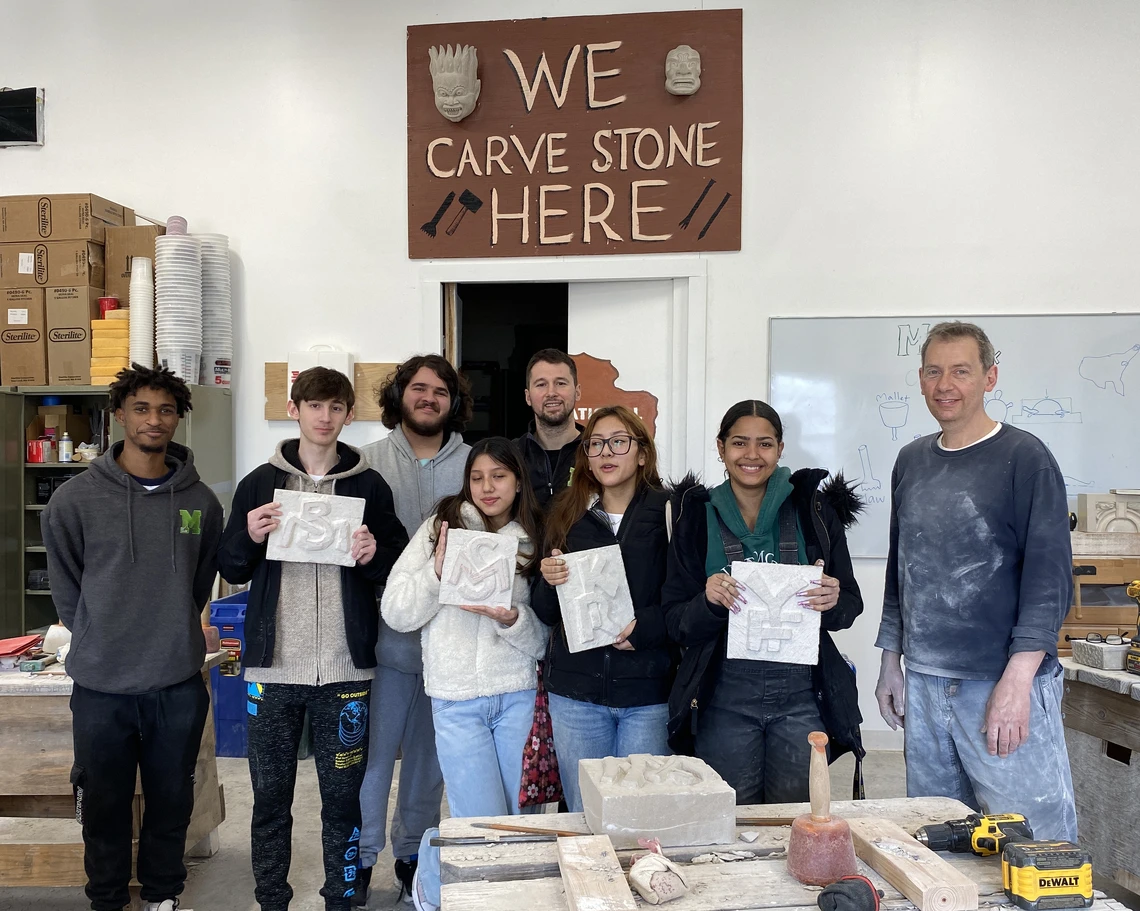 A group of seven high school students and their stone carving teacher pose in a workshop holding square limestone tablets that they carved with various designs; behind them a sign reads: “We Carve Stone Here.”