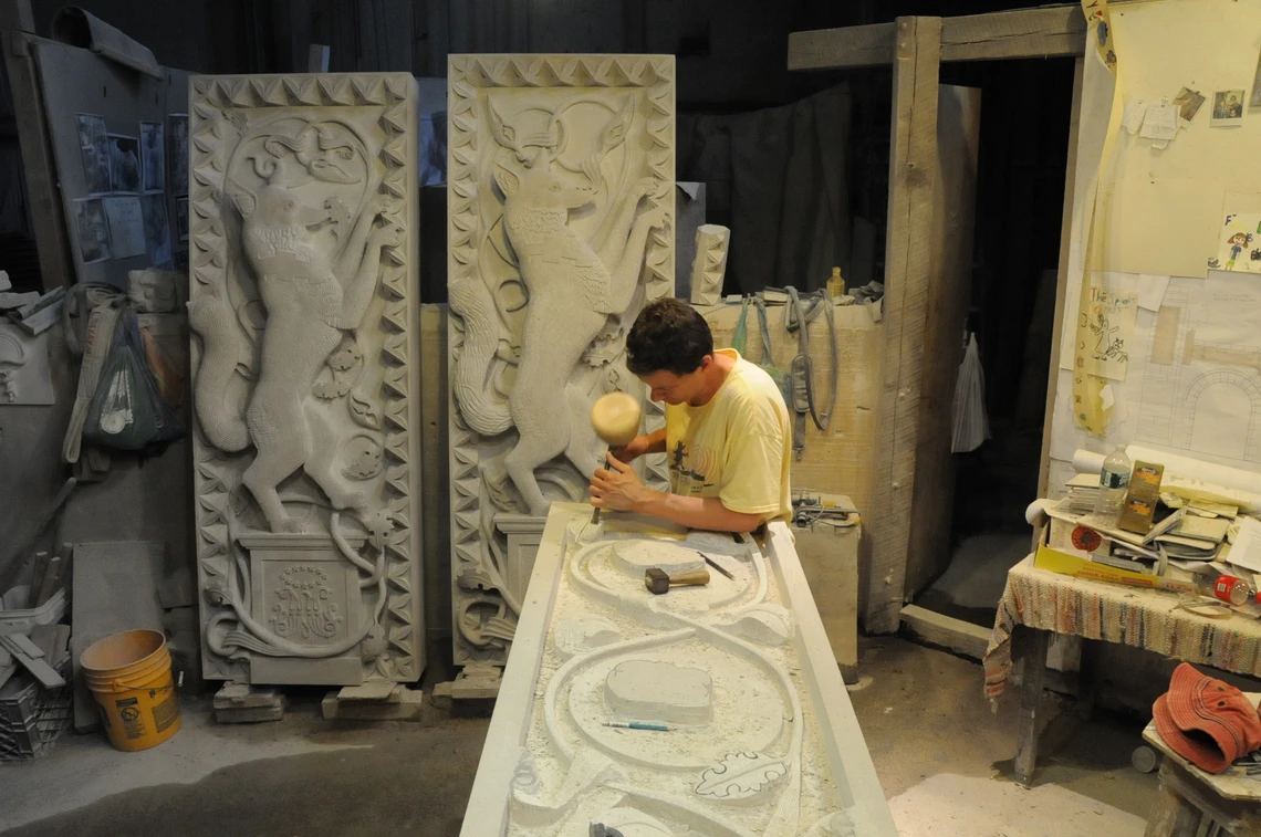 A man uses a mallet and chisel to carve a rectangular piece of limestone in a workshop; two large elaborate vertical carvings of a fox surrounded by ornamental motifs stand against the wall behind him.